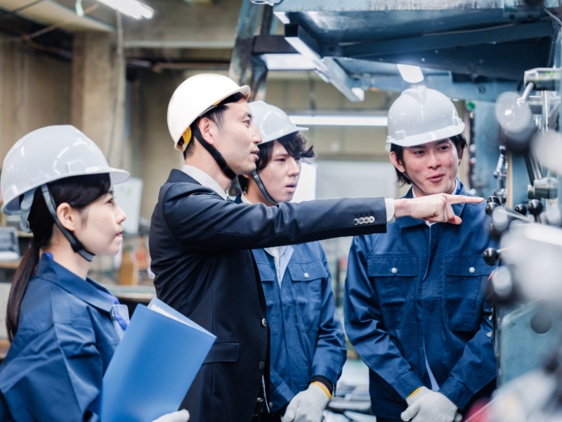 スーツ姿の上司がヘルメットを着用した若い作業員たちに機械の操作を指導している様子。